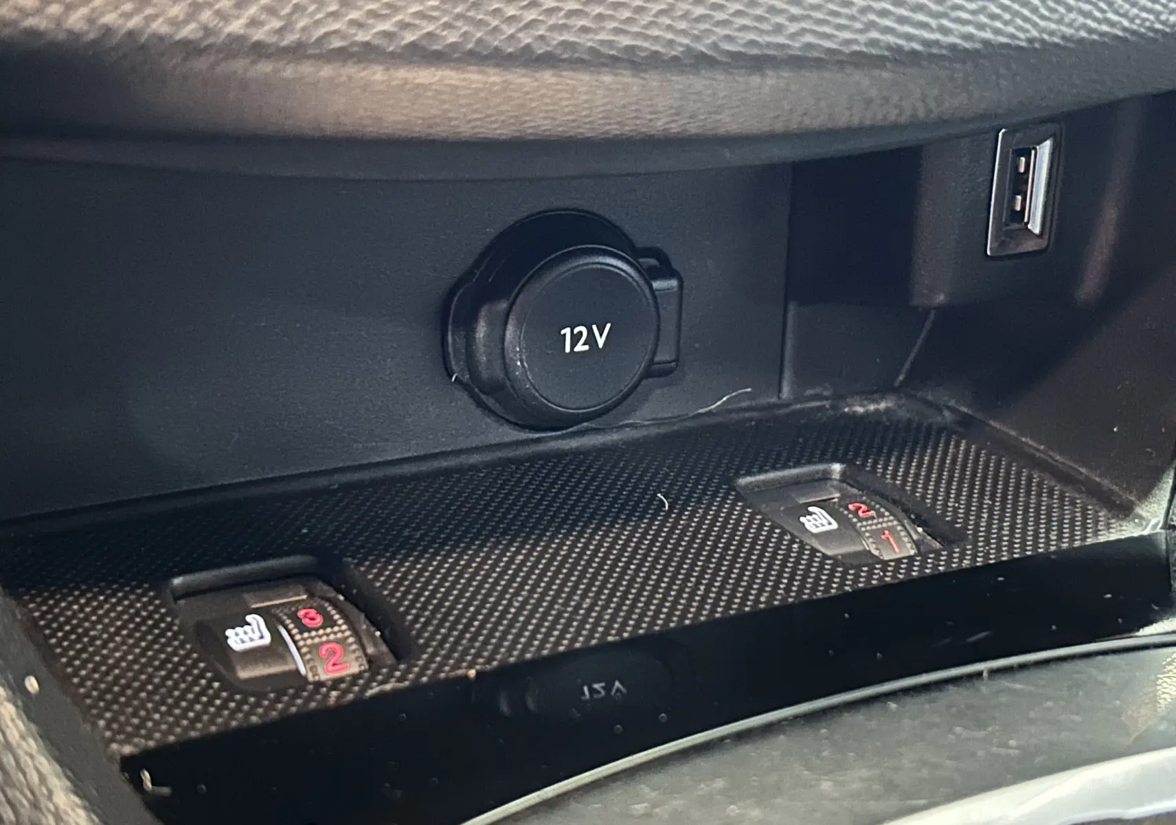 Gros plan sur la console centrale d'une Peugeot 308 gris, montrant les commandes de sièges chauffants et prise 12V avec port USB.