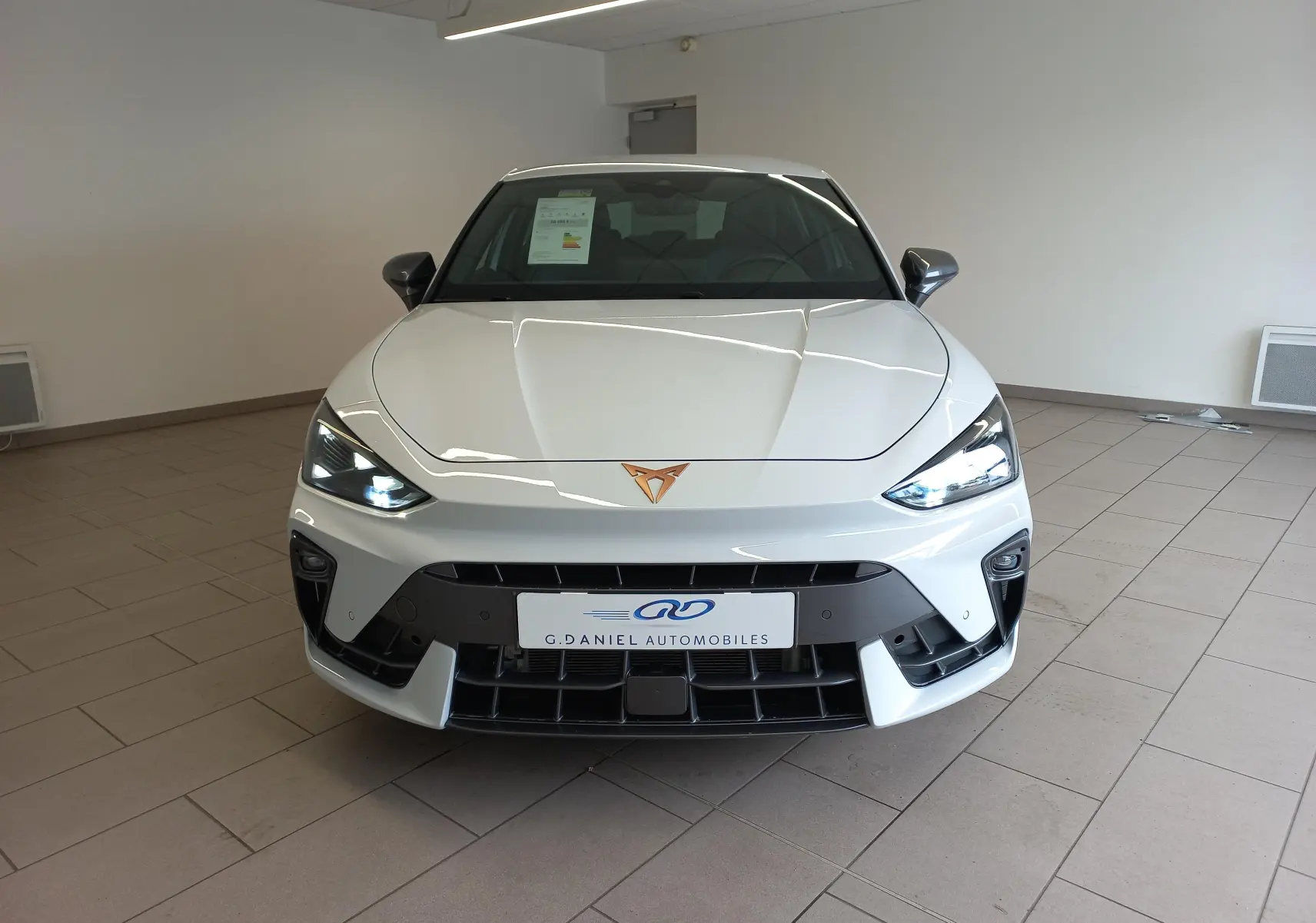 Vue frontale d'une CUPRA Leon Phase 2 blanche Nevada avec feux allumés et logo cuivre visible sur le capot.