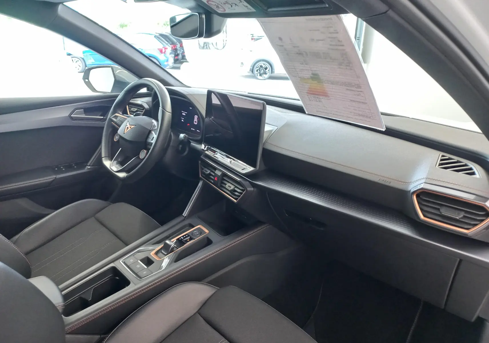 Vue intérieure côté conducteur de la CUPRA Leon 2025, tableau de bord noir avec écran tactile central et volant multifonction.