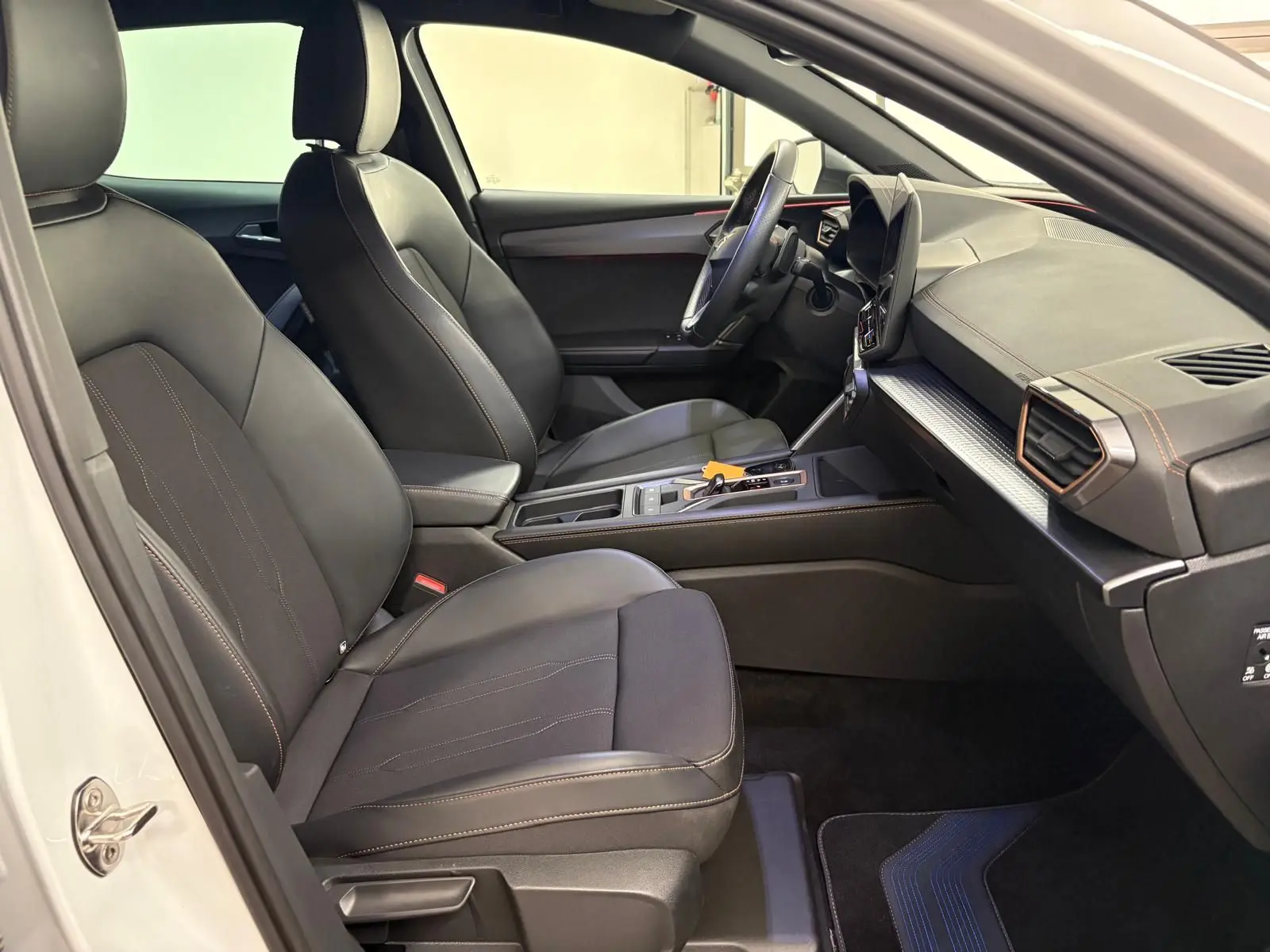 Vue intérieure côté conducteur de la CUPRA Leon blanc Nevada 2025 avec sièges noirs et surpiqûres orange.