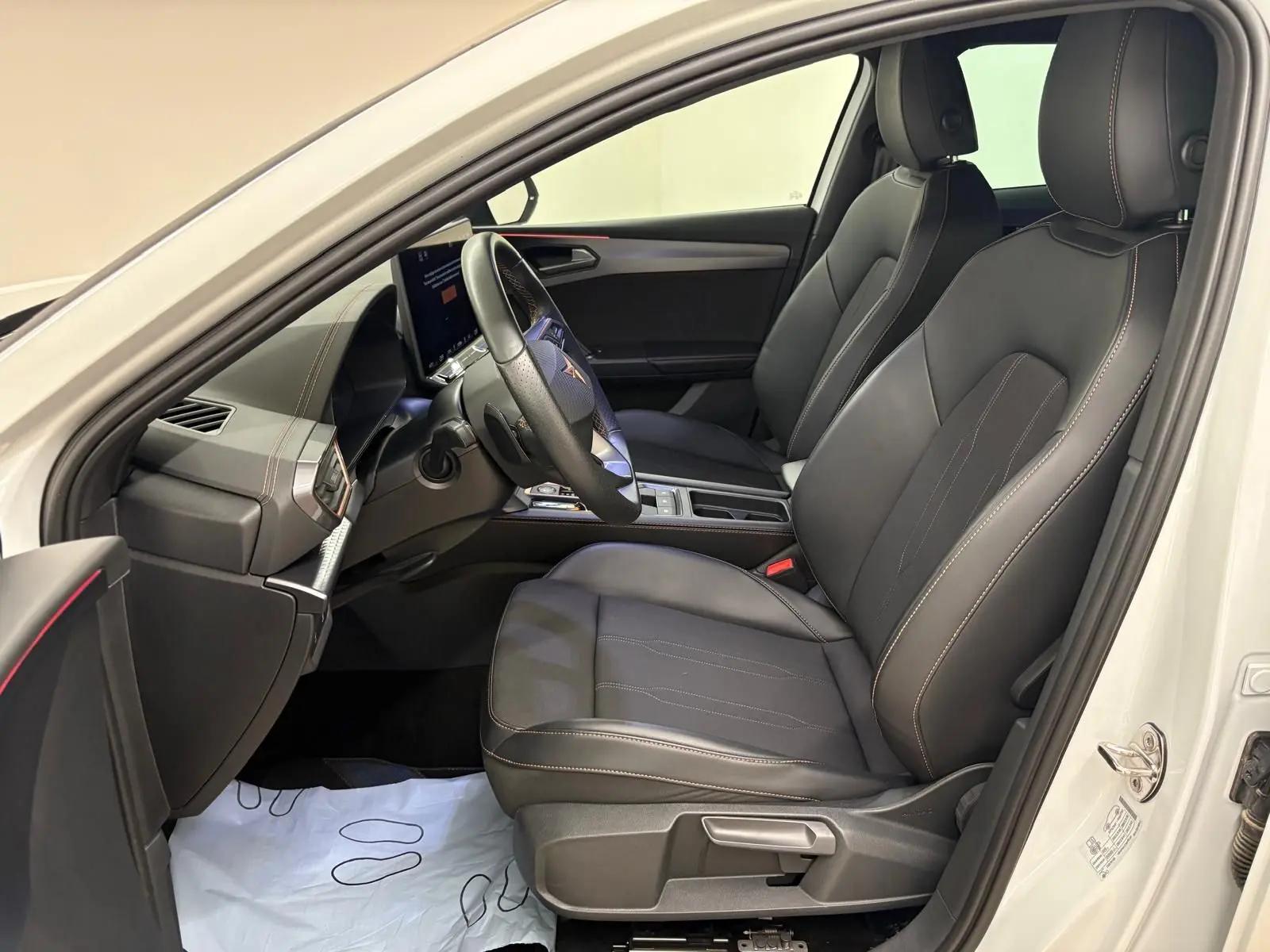 Vue intérieure côté conducteur de la CUPRA Leon blanc Nevada 2025, sièges noirs avec surpiqûres contrastantes et volant sport.