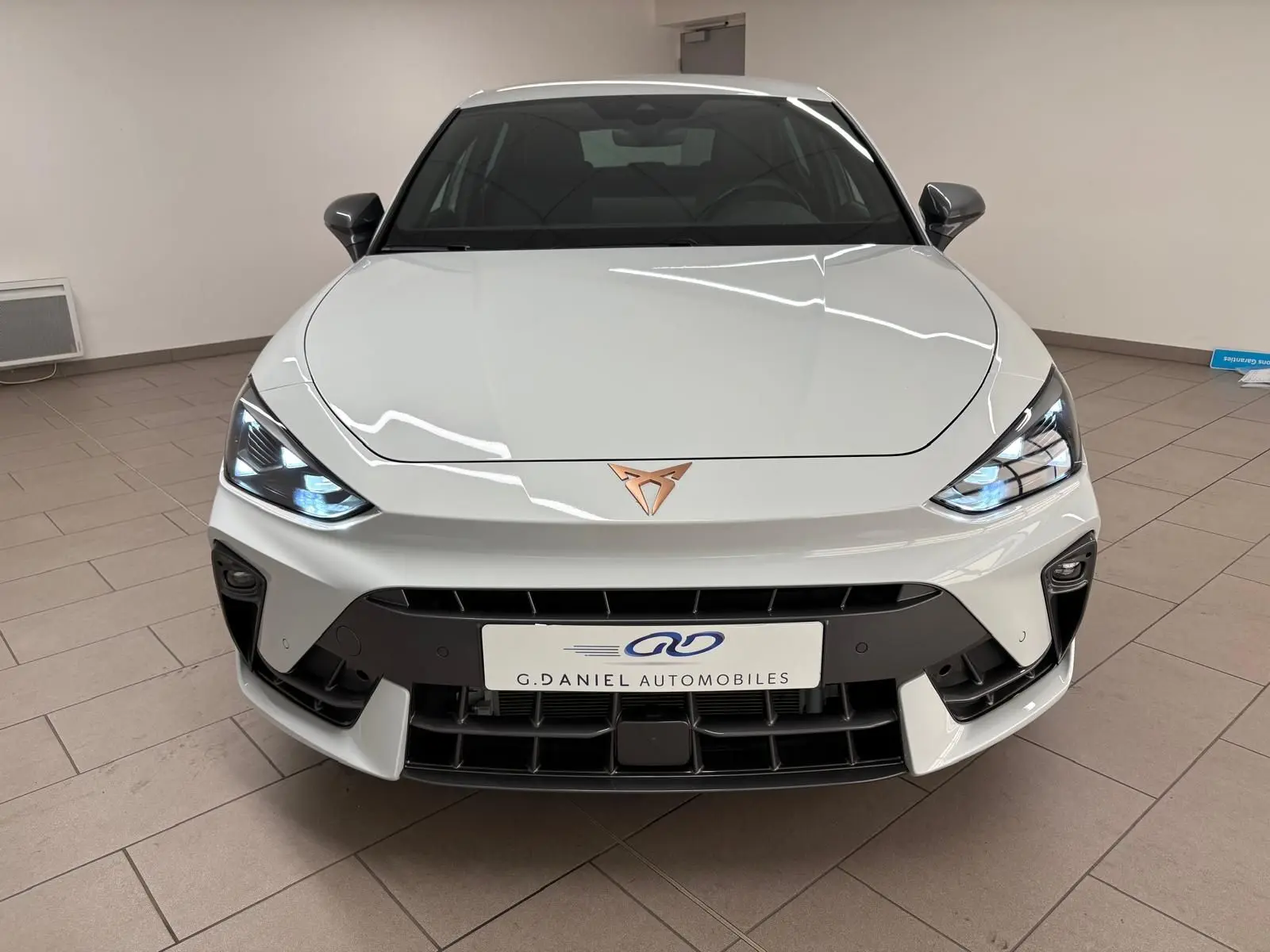 Vue frontale d'une CUPRA Leon Phase 2 blanche Nevada avec logo cuivré et phares allumés en intérieur.