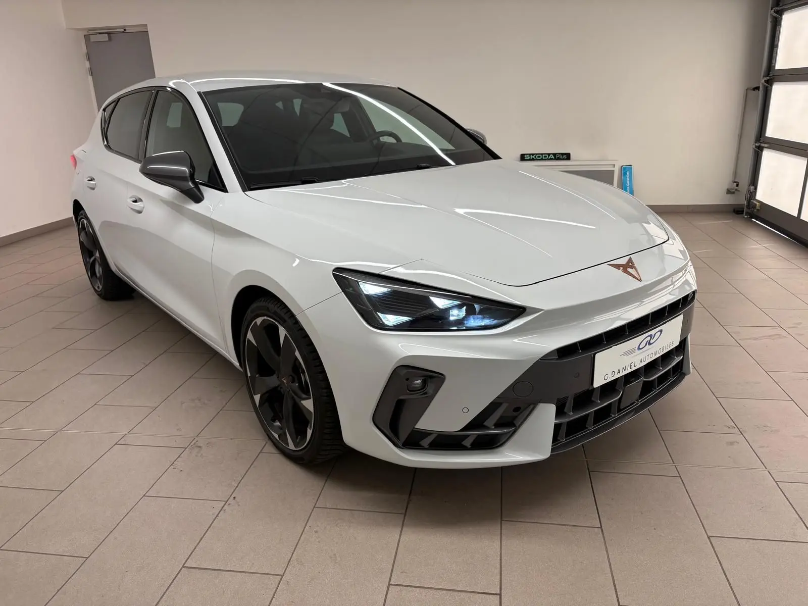 Vue 3/4 avant droite d'une CUPRA Leon blanc Nevada métallisé avec phares allumés et jantes noires distinctives.