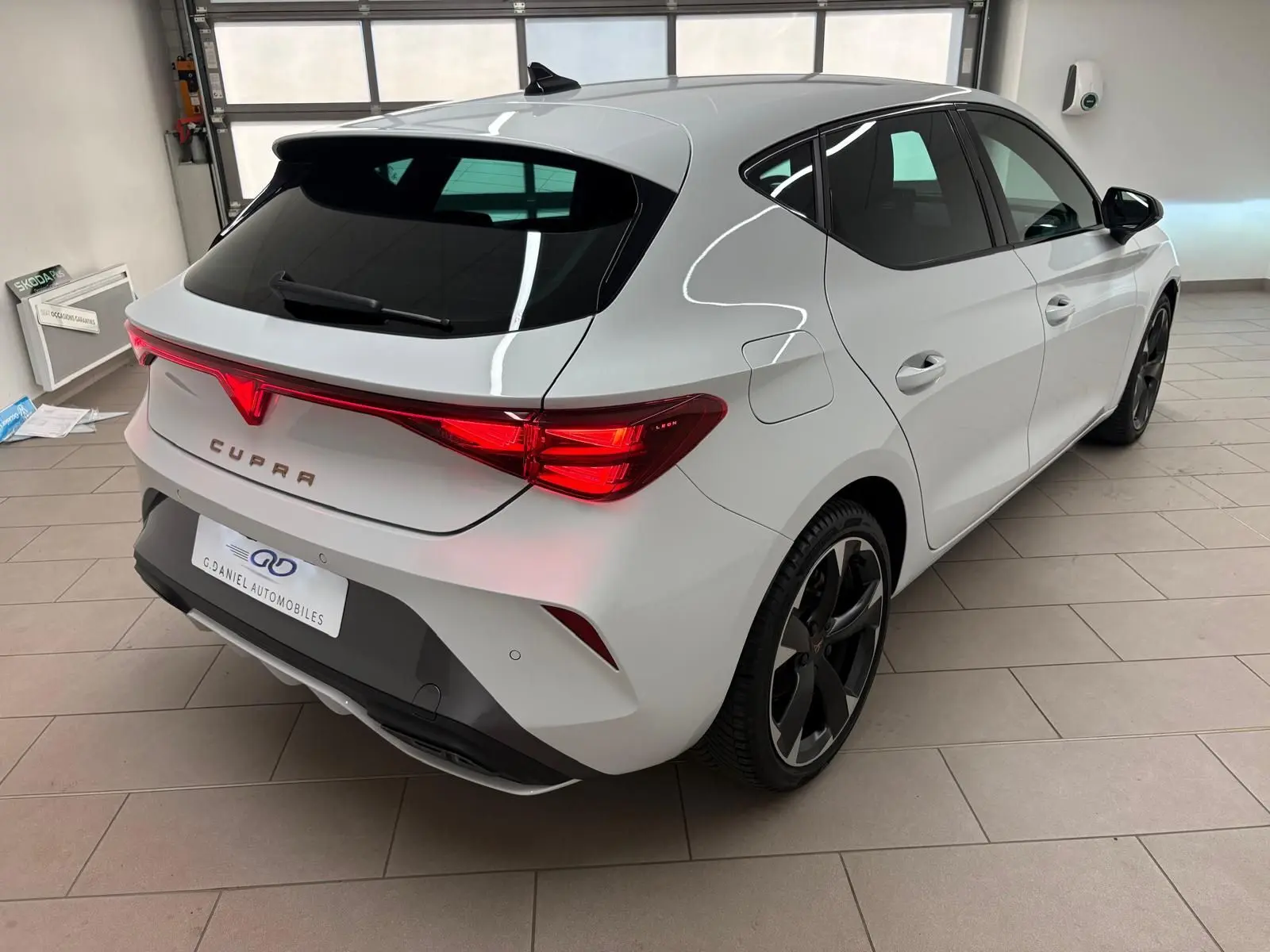 Vue 3/4 arrière droite d'une CUPRA Leon blanche Nevada avec feux arrière LED et jantes noires en intérieur showroom.