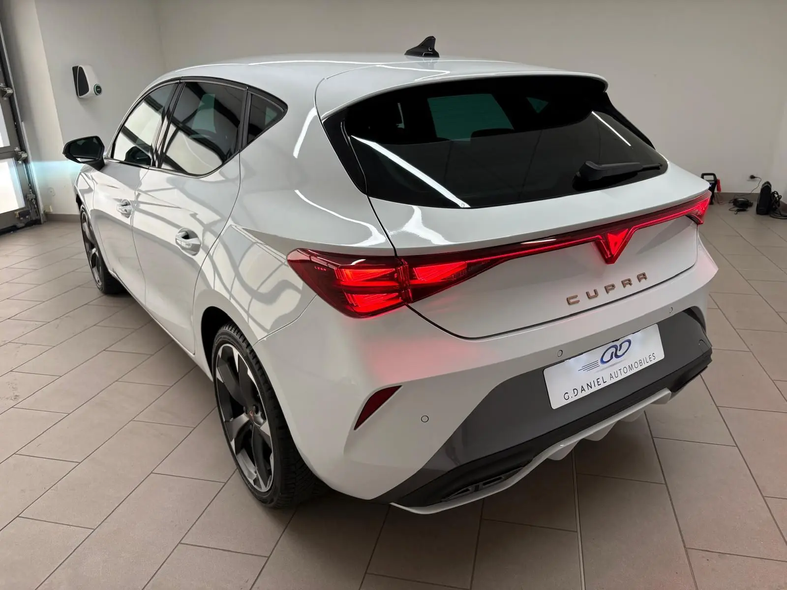 Vue 3/4 arrière droite d'une CUPRA Leon blanche Nevada avec feux arrière connectés et jantes noires en intérieur showroom.