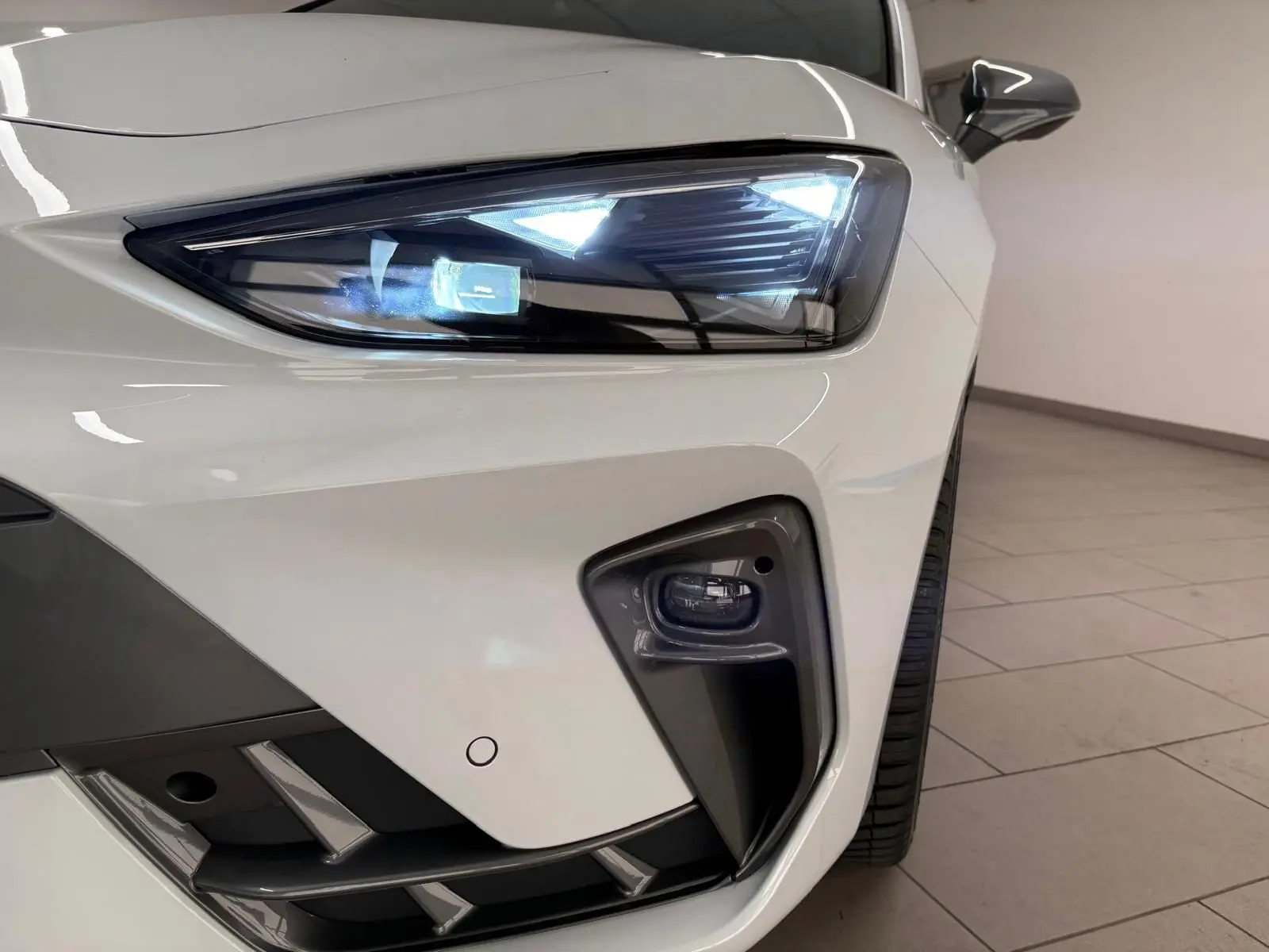 Gros plan sur le phare avant droit allumé d'une CUPRA Leon Phase 2 blanche Nevada avec antibrouillard intégré.