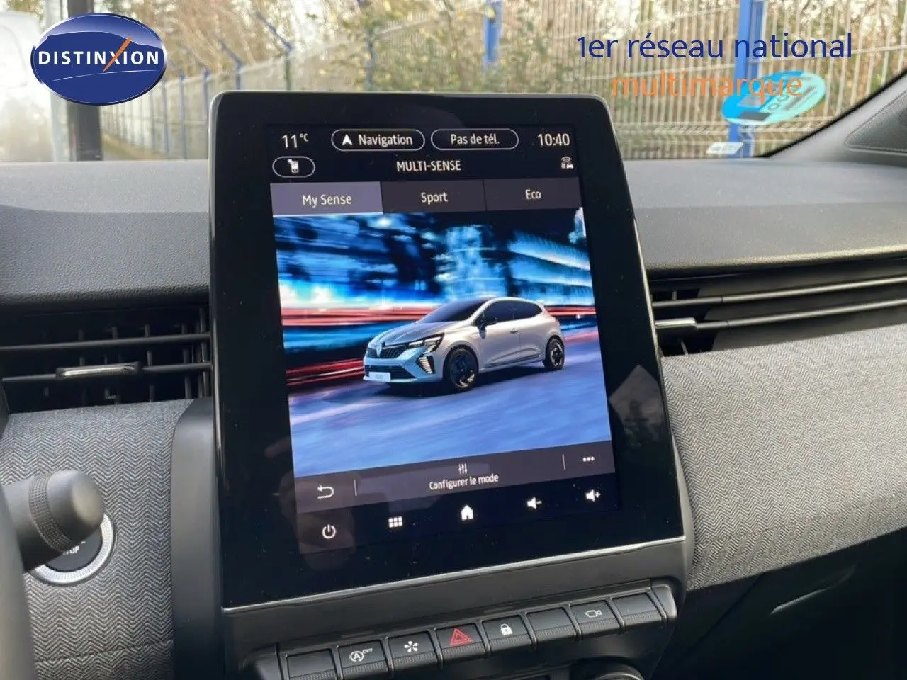 Écran tactile intérieur montrant une Renault Clio blanche en 3/4 avant dans un décor flouté urbain.