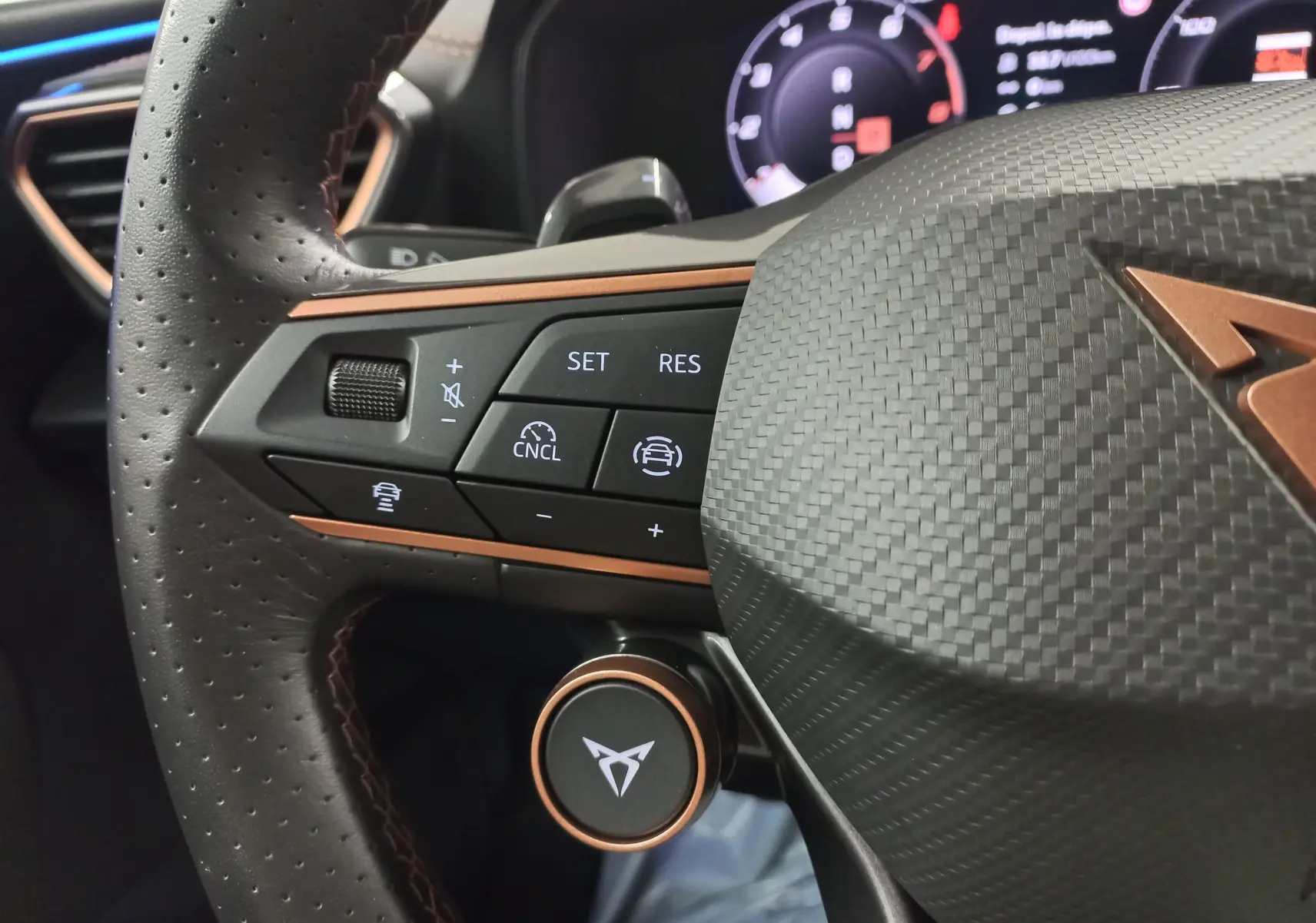 Gros plan sur le volant cuir perforé et carbone du CUPRA Leon Phase 2 avec commandes intégrées et logo cuivre.