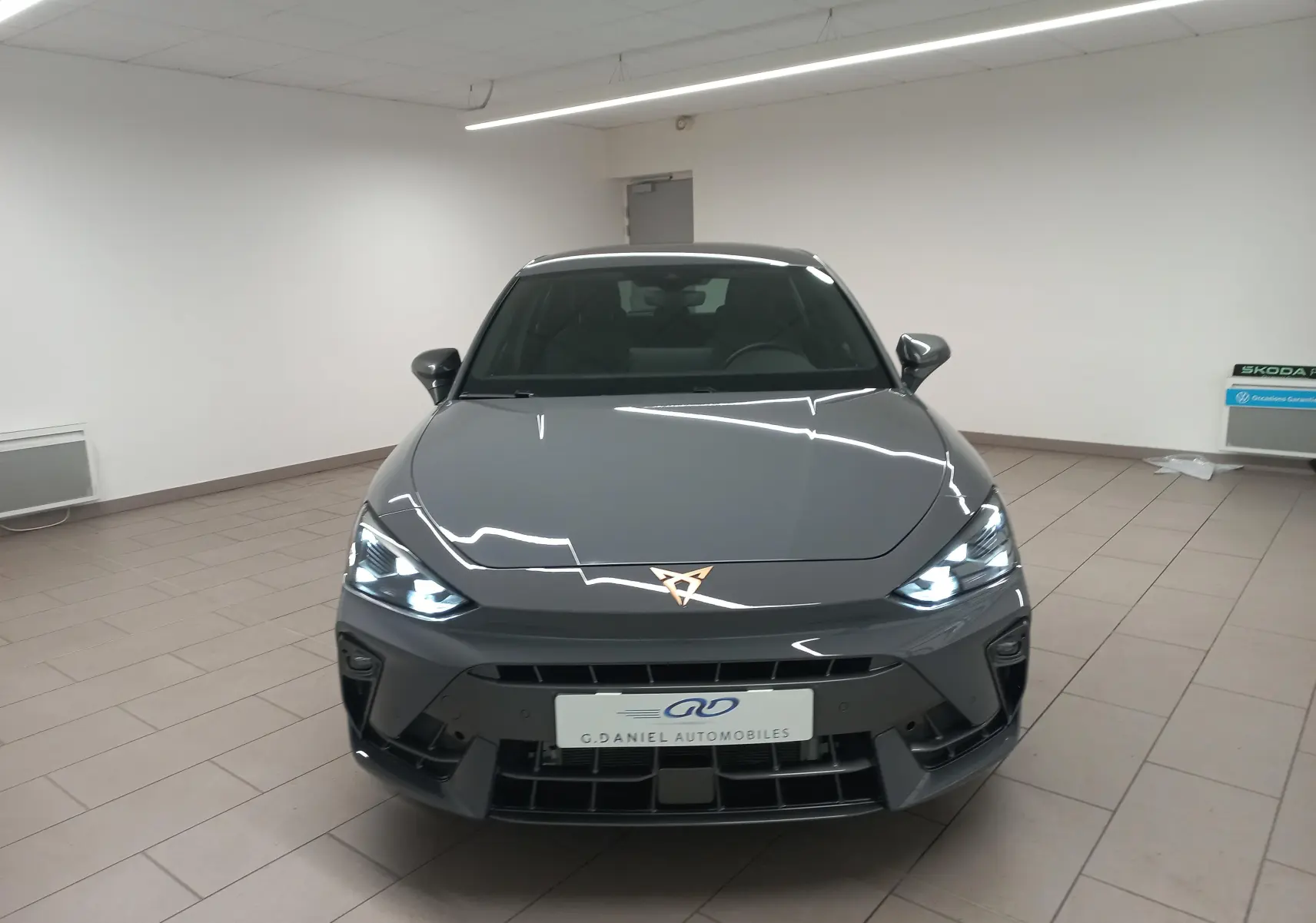 Vue avant d'une CUPRA Leon Phase 2 gris Graphene avec phares allumés et logo CUPRA lumineux au centre du capot.