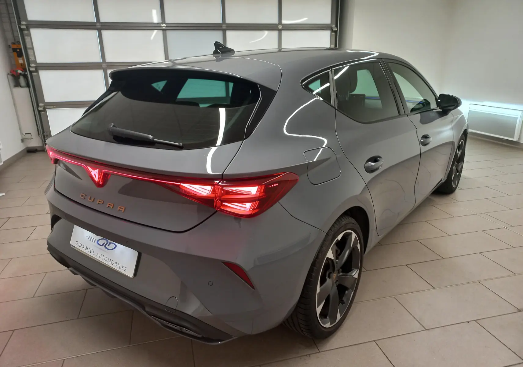 Vue 3/4 arrière droite d'une CUPRA Leon 2025 gris Graphene avec feux arrière allumés et jantes noires en intérieur showroom.