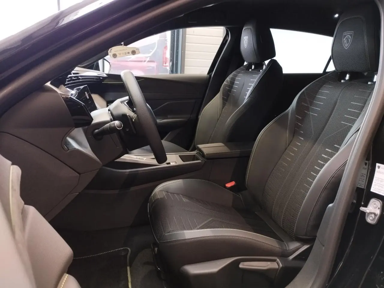 Vue intérieure côté conducteur de la Peugeot 308 Hybrid 2026 noire, sièges avec logo Lion et surpiqûres vert Adamite.