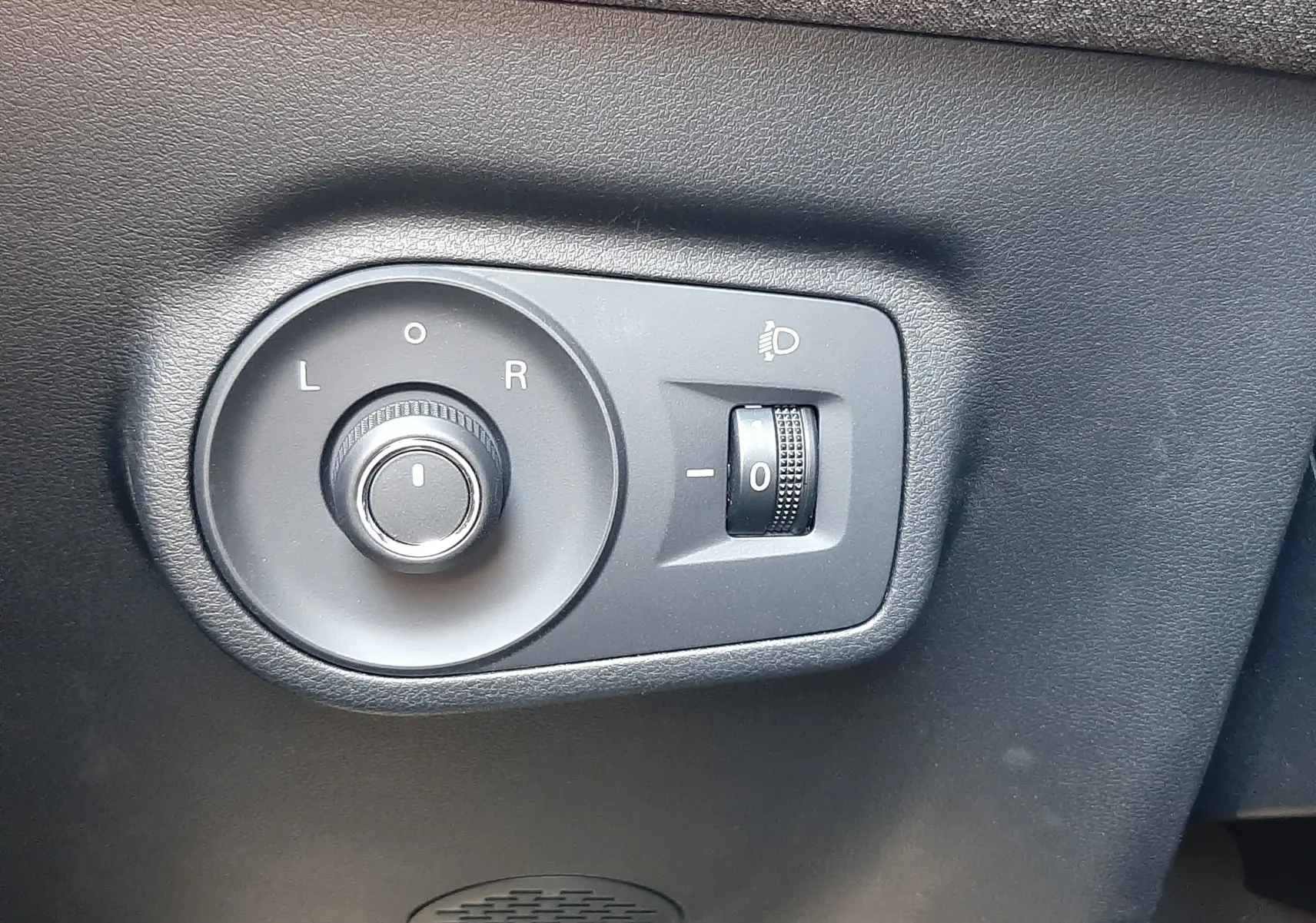 Gros plan sur les commandes de réglage des rétroviseurs et des phares, sur panneau noir intérieur d'un MG Motor ZS Pebble Black.