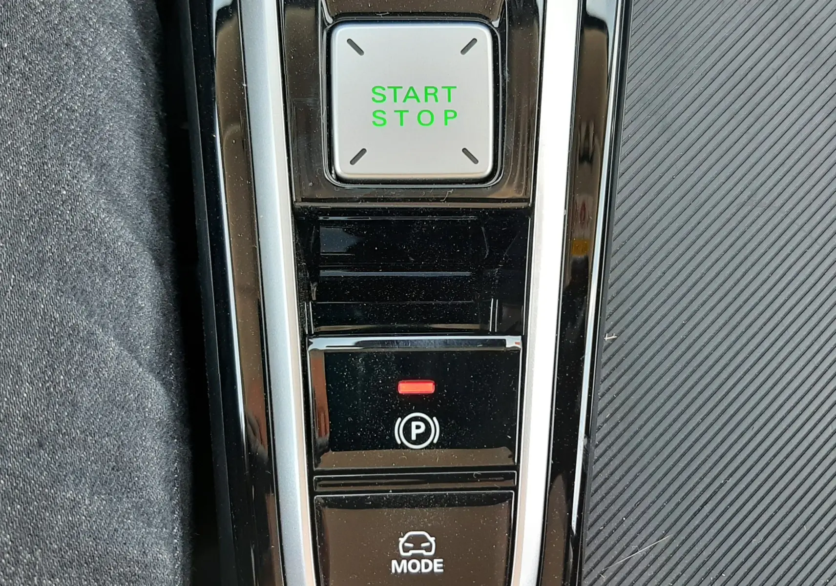 Gros plan sur la console centrale noire brillante du MG ZS 1.5L Hybrid+ avec bouton Start/Stop vert et commandes de frein à main et mode de conduite.