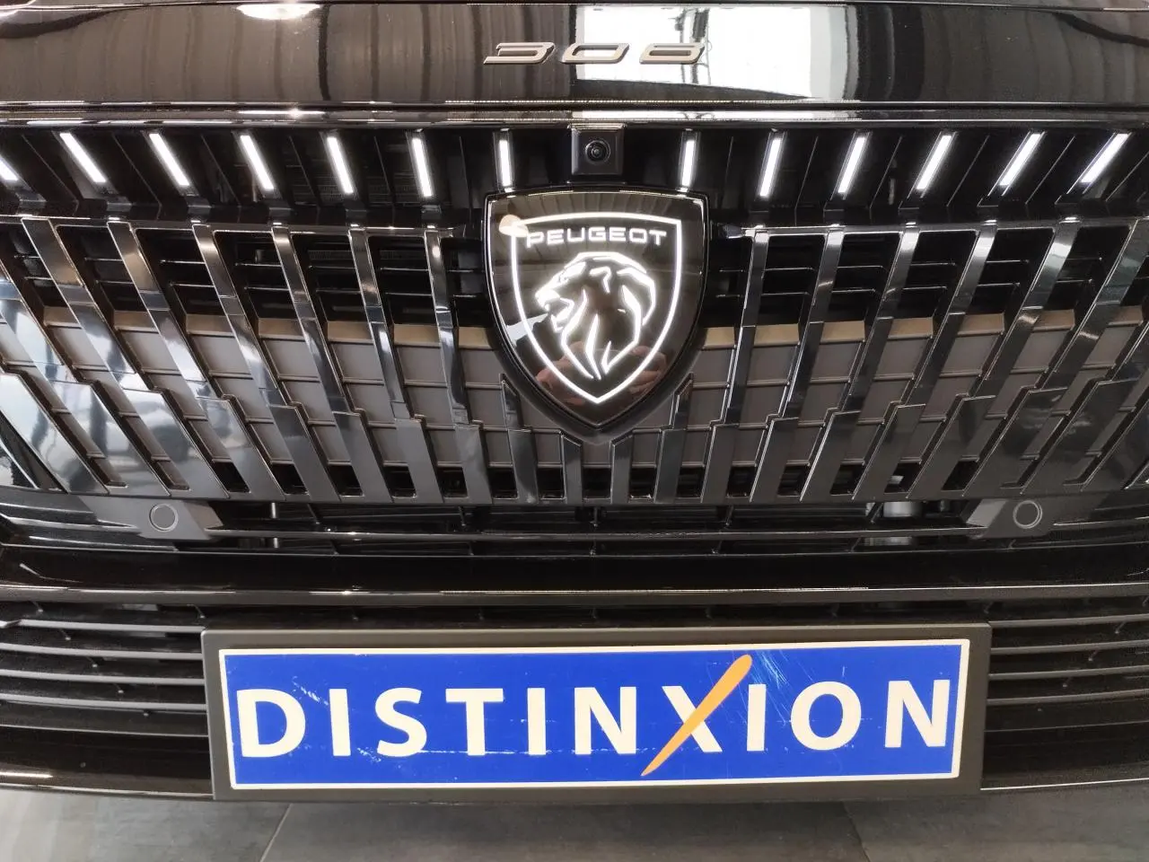 Gros plan sur la calandre noire métallisée de la Peugeot 308 Hybrid 2026 avec logo Peugeot illuminé et plaque Distinxion.