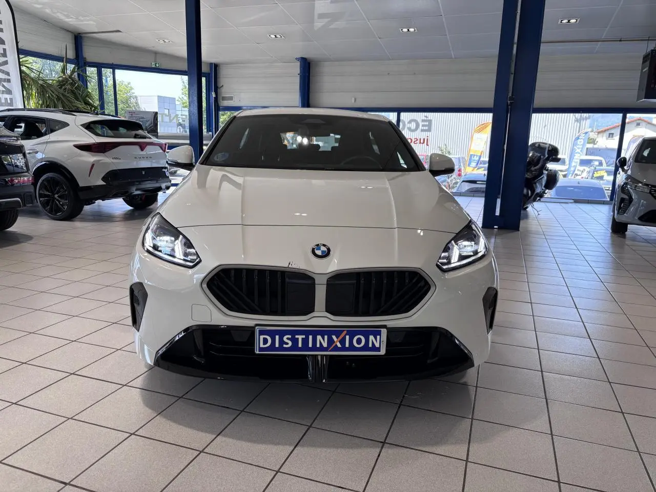 Vue avant d'une BMW Série 1 120d M Sport blanche 2025 en showroom, avec calandre noire et phares allumés.
