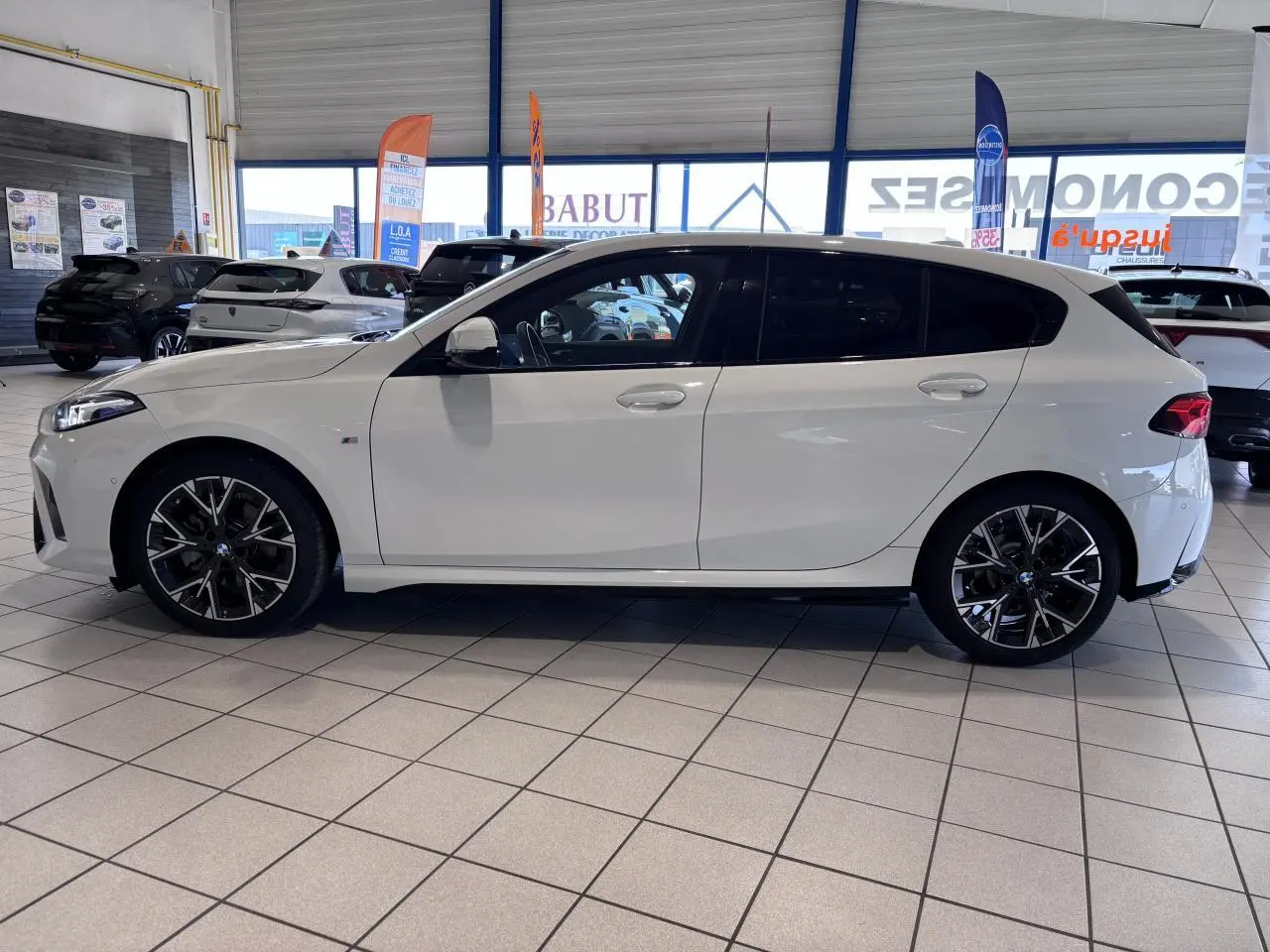 BMW Série 1 120d M Sport blanc vue de profil côté gauche, avec jantes noires et vitres teintées, en showroom.