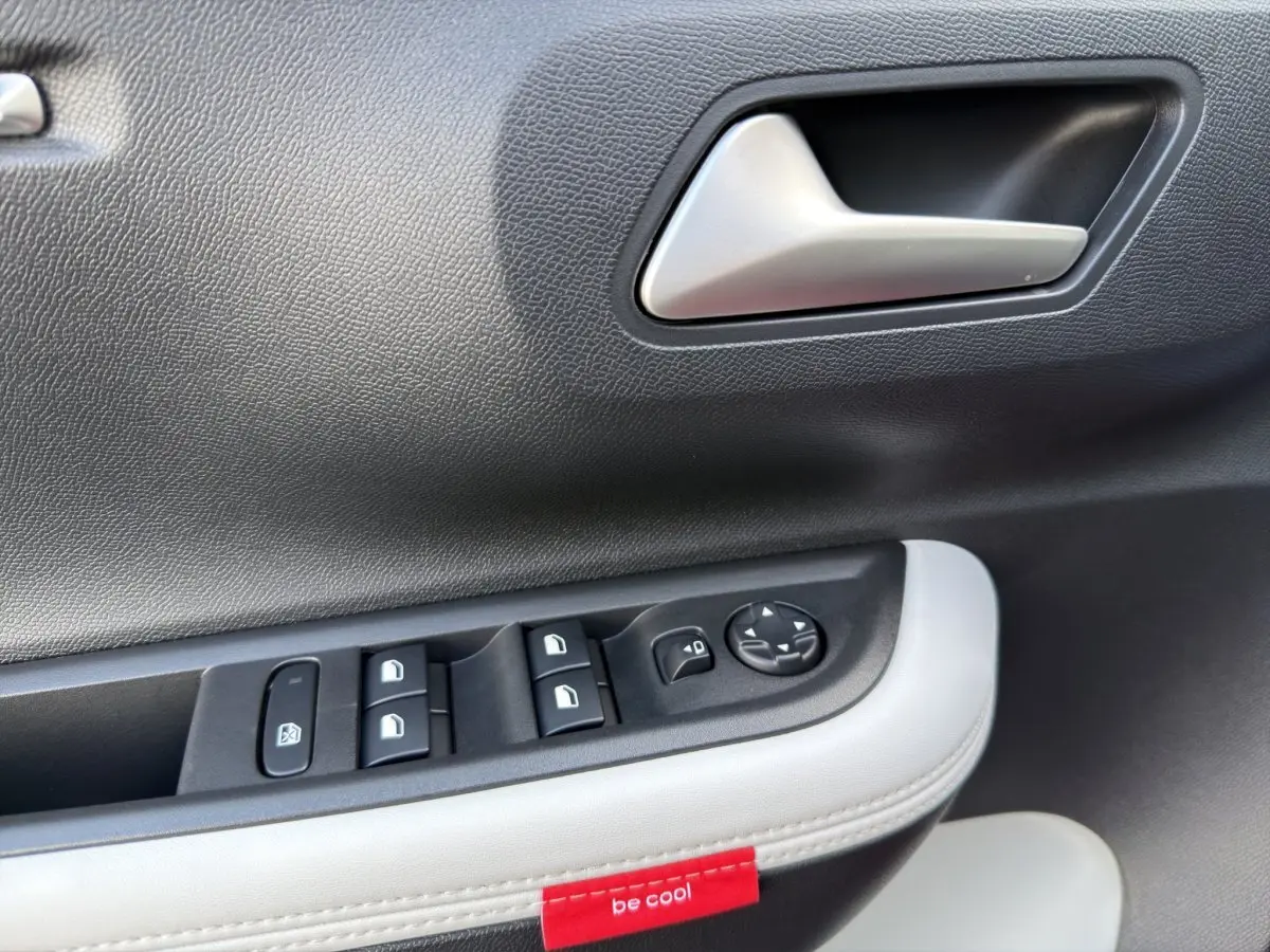 Détail de la porte côté conducteur avec commandes électriques et étiquette rouge "be cool" sur un intérieur noir et blanc.
