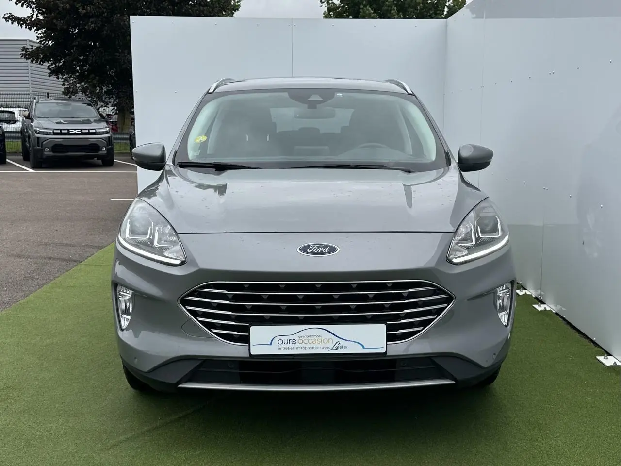 Vue de face d'un Ford Kuga gris solaire avec calandre chromée et feux LED allumés sur un sol vert.