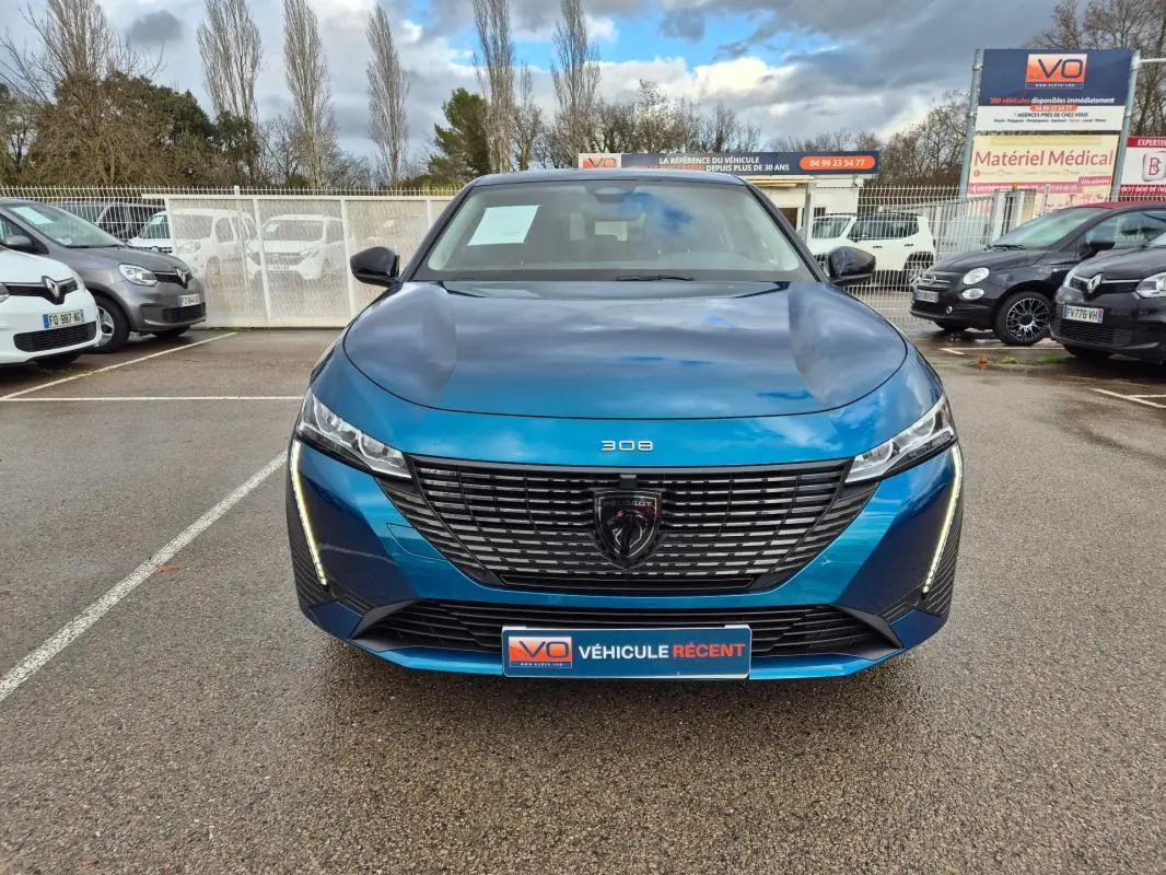 Vue frontale d'une Peugeot 308 bleu métallisé 2024 avec calandre noire et feux LED verticaux allumés.