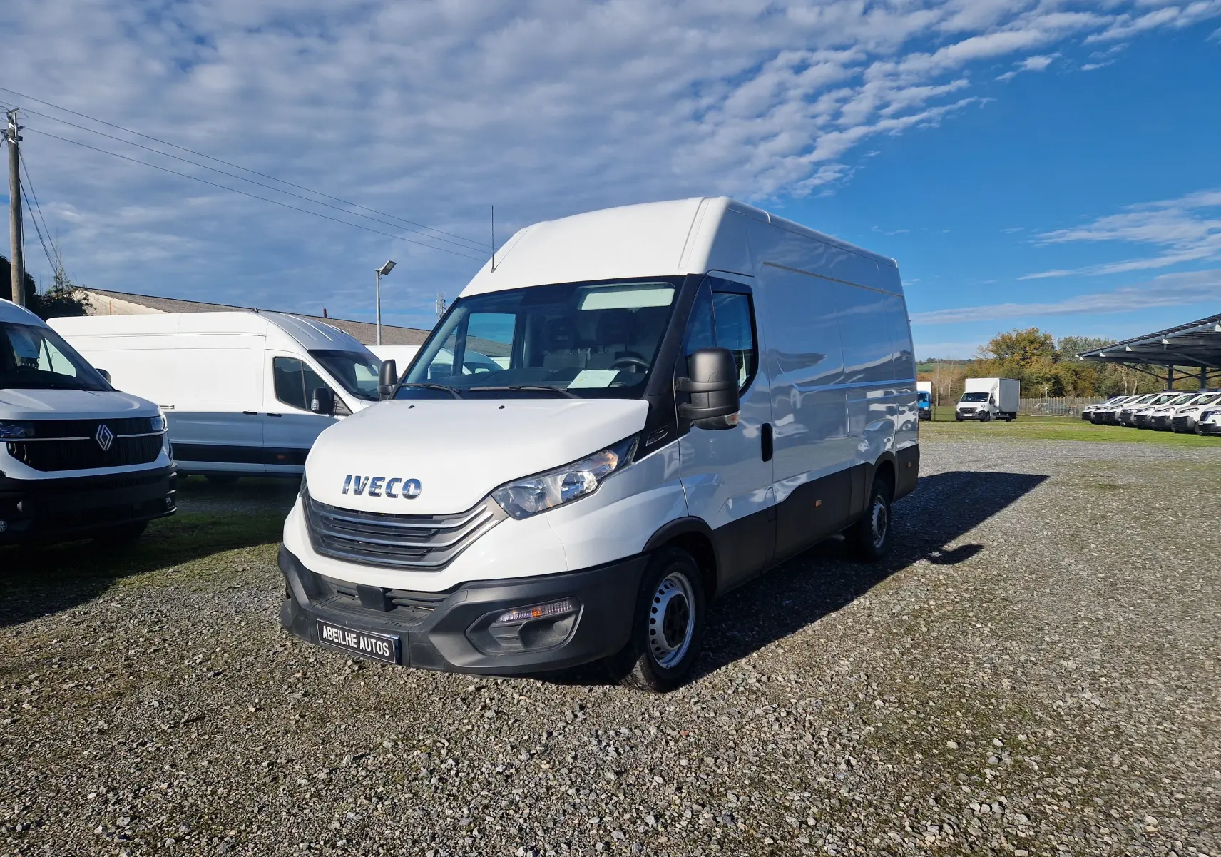 Fourgon blanc Iveco Daily 2022 vu en 3/4 avant droit sur un terrain gravillonné avec plusieurs autres véhicules en arrière-plan.