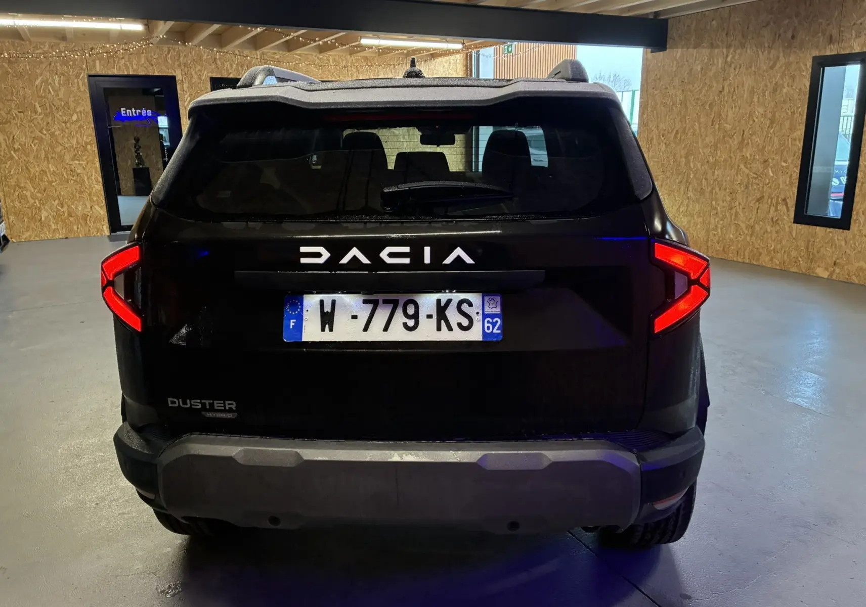 Vue arrière d'un Dacia Duster 1.6 Hybrid 140 noir, avec feux arrière allumés dans un garage.