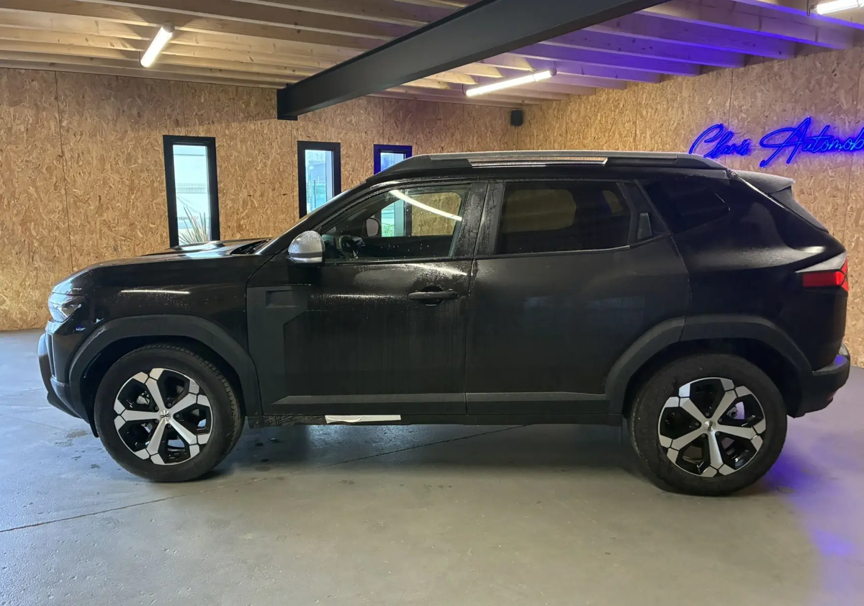 Vue de profil côté gauche d'un Dacia Duster 1.6 Hybrid 140 Journey noir avec jantes bicolores en intérieur industriel.