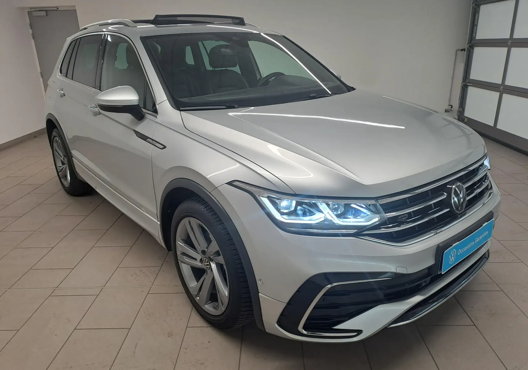 Volkswagen Tiguan 2024 finition R-Line en Reflet d'Argent, vue 3/4 avant avec phares allumés dans un garage.
