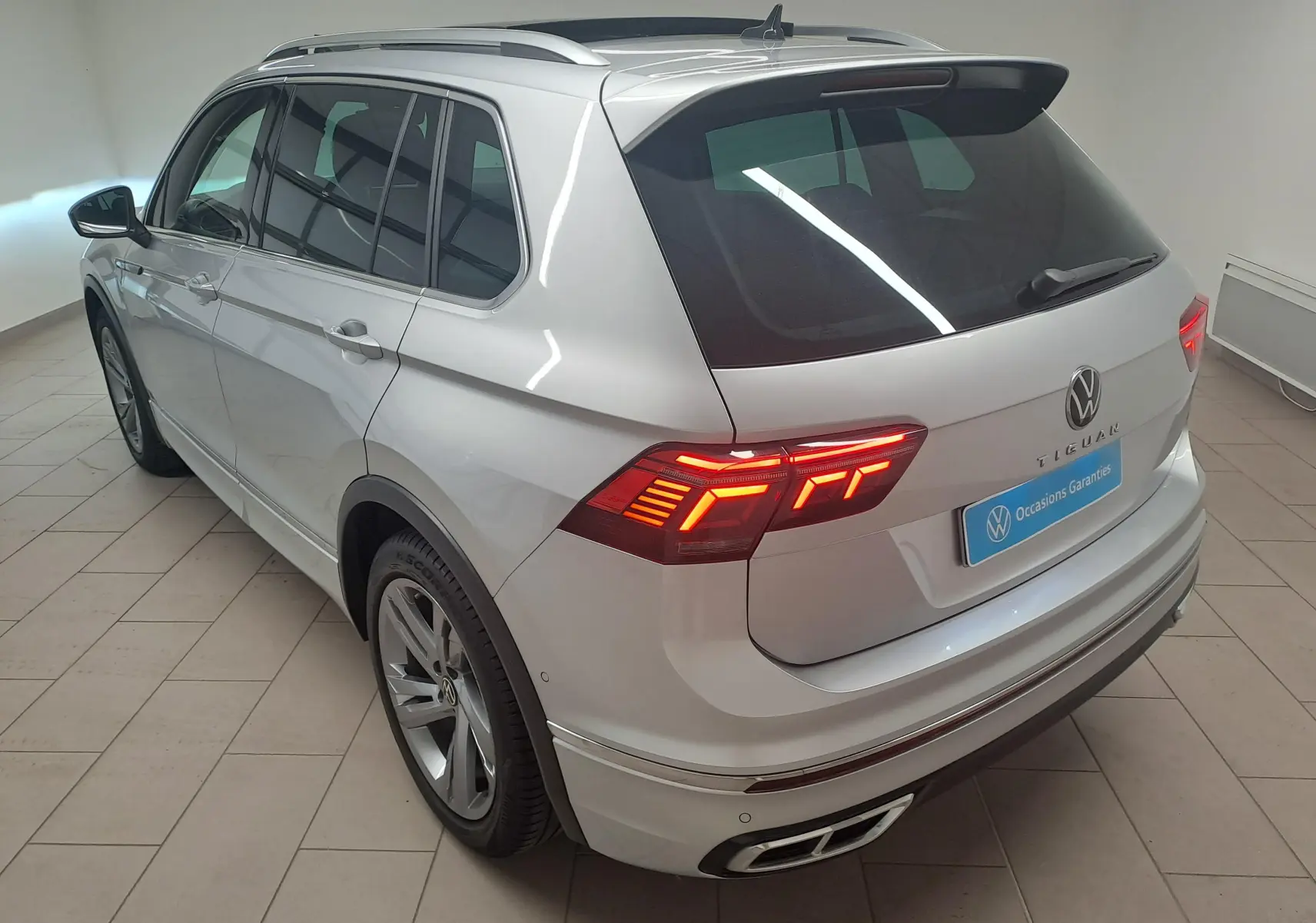 Volkswagen Tiguan 2024 finition R-Line en Reflet d'Argent, vue 3/4 arrière droit avec feux LED allumés et attelage escamotable.