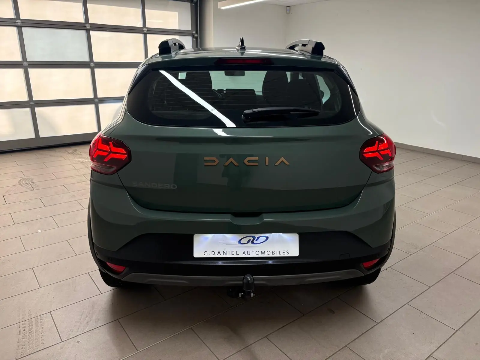 Vue arrière d'une Dacia Sandero Stepway 2023 vert cèdre avec feux allumés et attelage visible en intérieur.