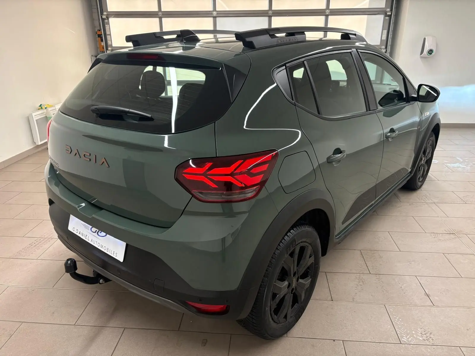 Vue 3/4 arrière droite du Dacia Sandero Stepway 2023 vert cèdre avec attelage et jantes noires en intérieur showroom.