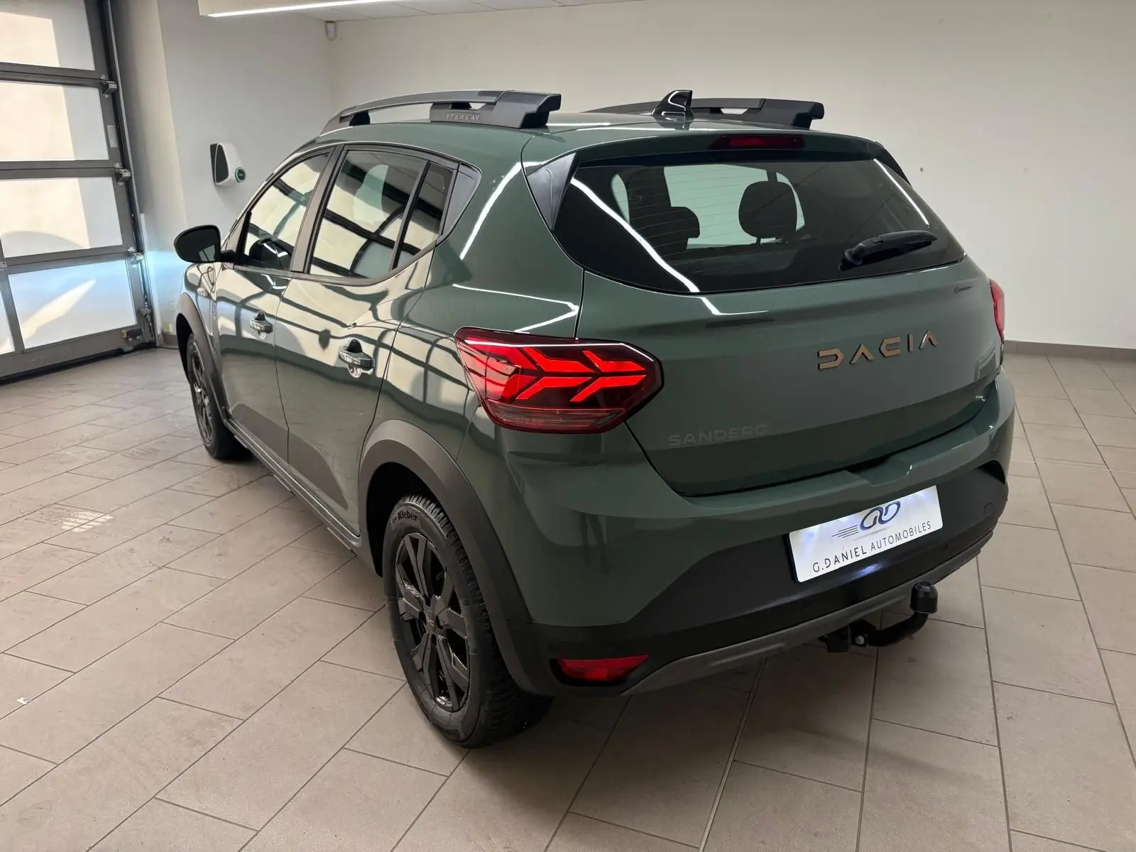 Vue 3/4 arrière droite d'une Dacia Sandero Stepway 2023 vert cèdre avec attelage et jantes noires en intérieur showroom.
