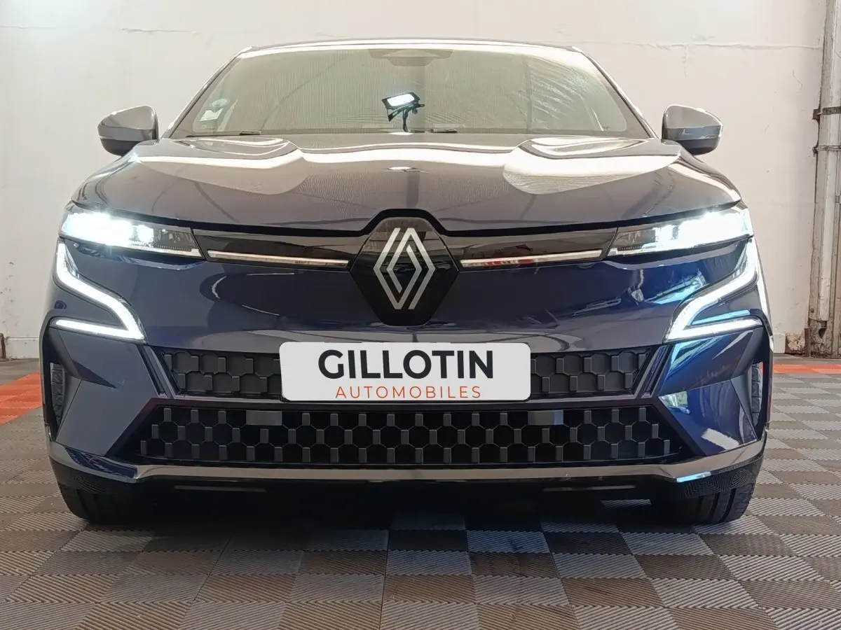 Vue frontale d'une Renault Mégane Techno EV60 bleu avec feux LED allumés et calandre noire à motifs géométriques.