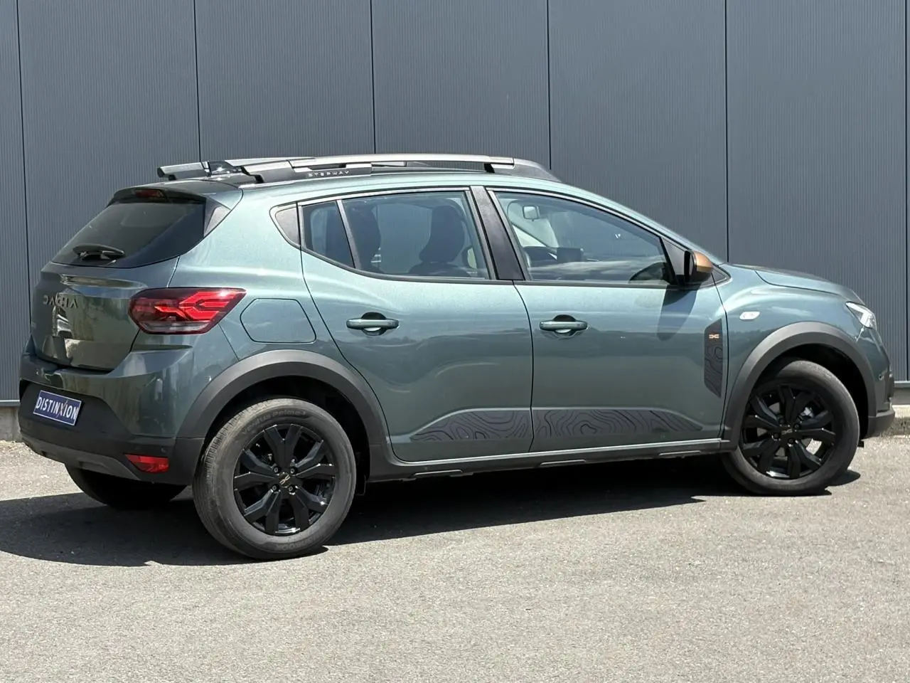 Vue de profil côté droit d'une Dacia Sandero Stepway Vert Cèdre avec jantes noires et barres de toit gris Mégalithe.