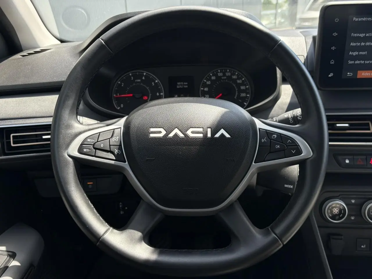 Vue frontale du volant noir DACIA avec commandes intégrées et tableau de bord numérique en arrière-plan.