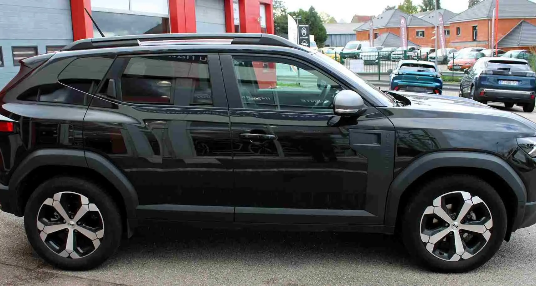 Profil droit d'un Dacia Duster noir nacré 2025 avec jantes alliage 18 pouces et barres de toit noires.