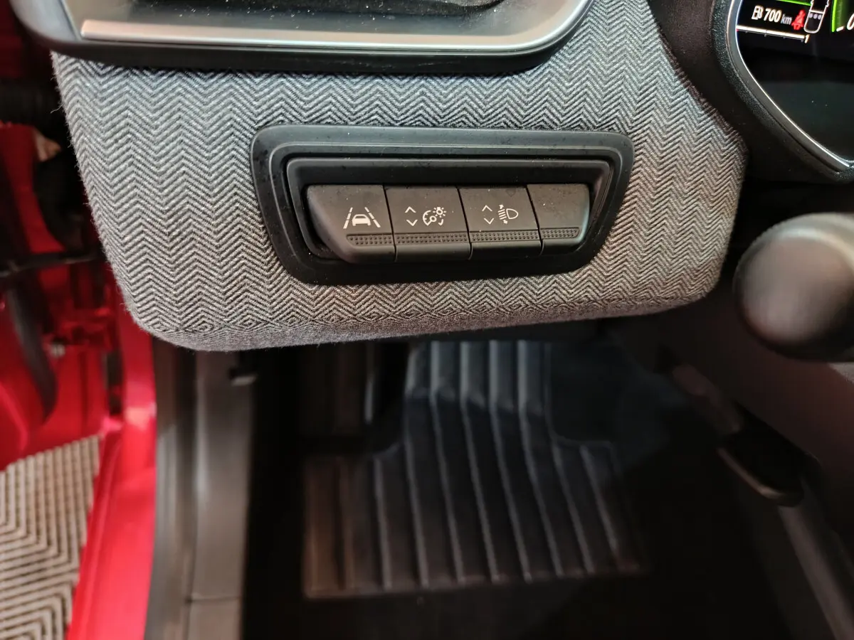 Gros plan sur les commandes d'aide à la conduite sous le tableau de bord, avec habillage gris et portière rouge visible côté gauche.