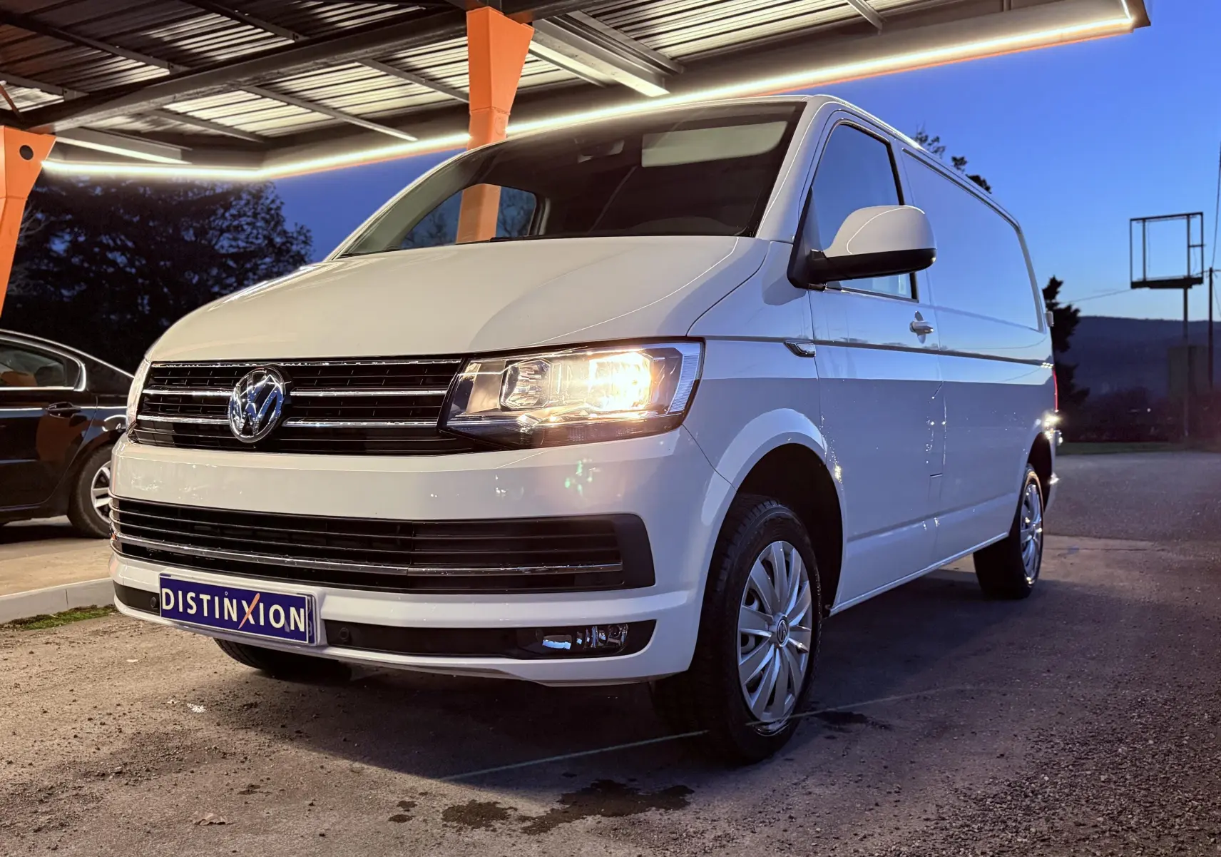 Volkswagen Transporter L1H1 blanc vu en 3/4 avant droit, phare allumé, stationné sous un abri au crépuscule.