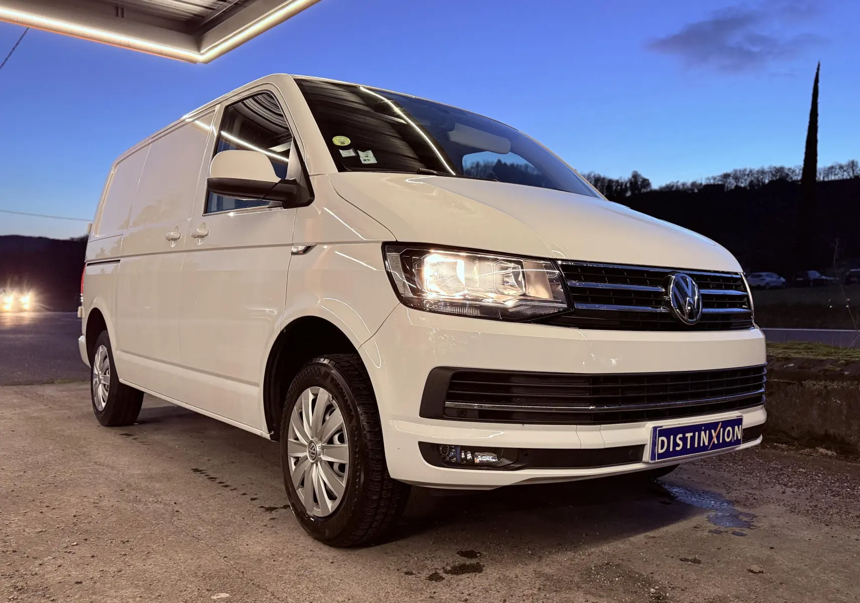 Volkswagen Transporter L1H1 blanc vu en 3/4 avant droit avec phares allumés au crépuscule.