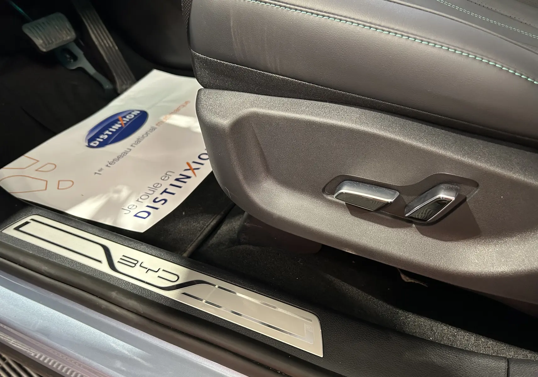 Gros plan sur les commandes de réglage du siège côté gauche et la plaque de seuil BYD dans l'habitacle noir.