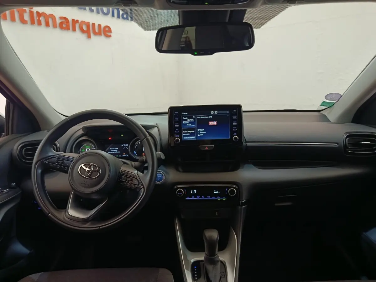 Vue intérieure frontale du tableau de bord noir de la Toyota Yaris Hybride 116h avec écran tactile central et volant cuir multifonctions.
