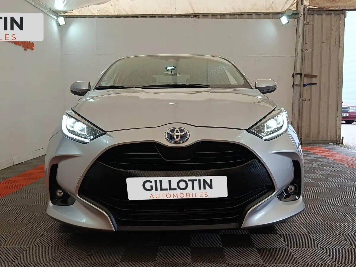 Vue frontale d'une Toyota Yaris Hybride 116h Design gris clair avec phares LED allumés dans un showroom.