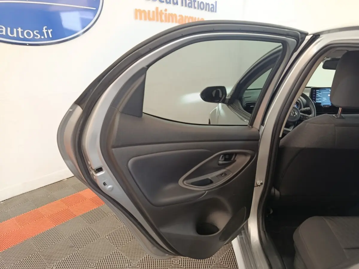Vue intérieure de la porte arrière gauche ouverte d'une Toyota Yaris hybride gris clair, avec siège arrière et tableau de bord partiellement visibles.