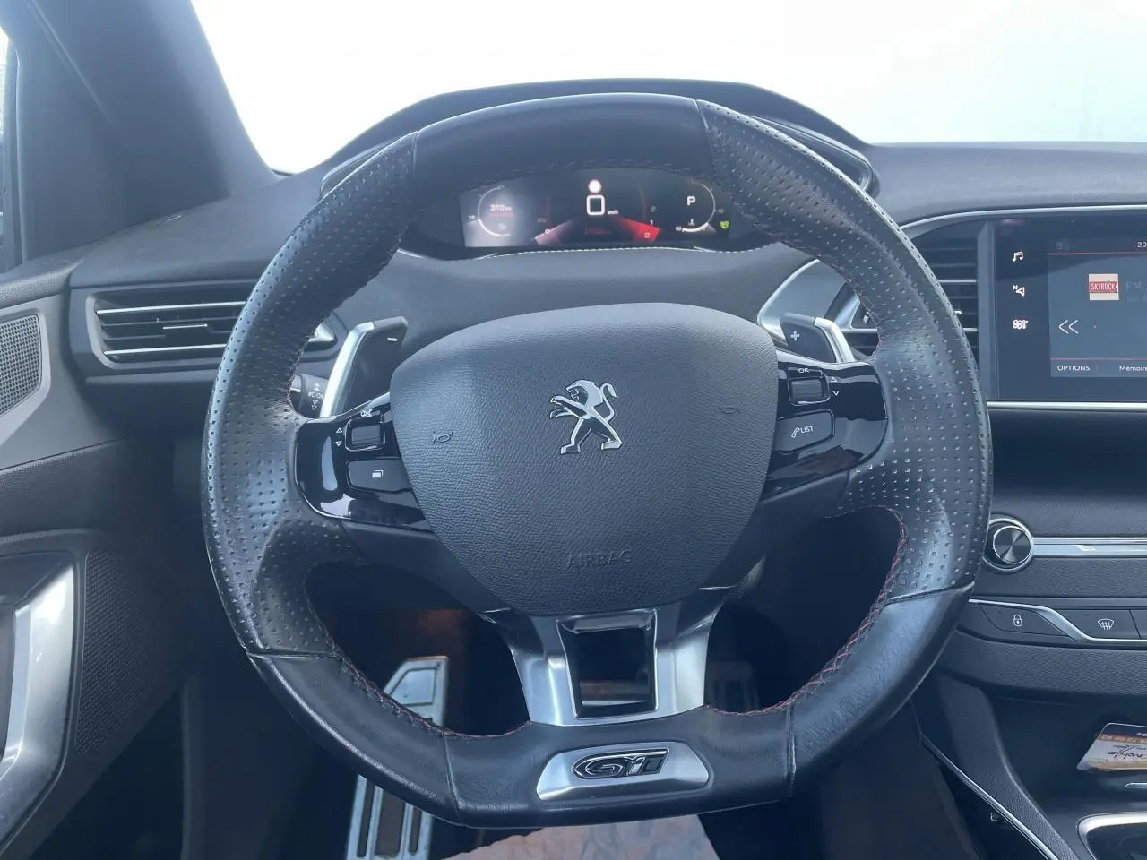 Vue rapprochée du volant cuir perforé noir avec logo Peugeot et badge GT du tableau de bord intérieur anthracite.