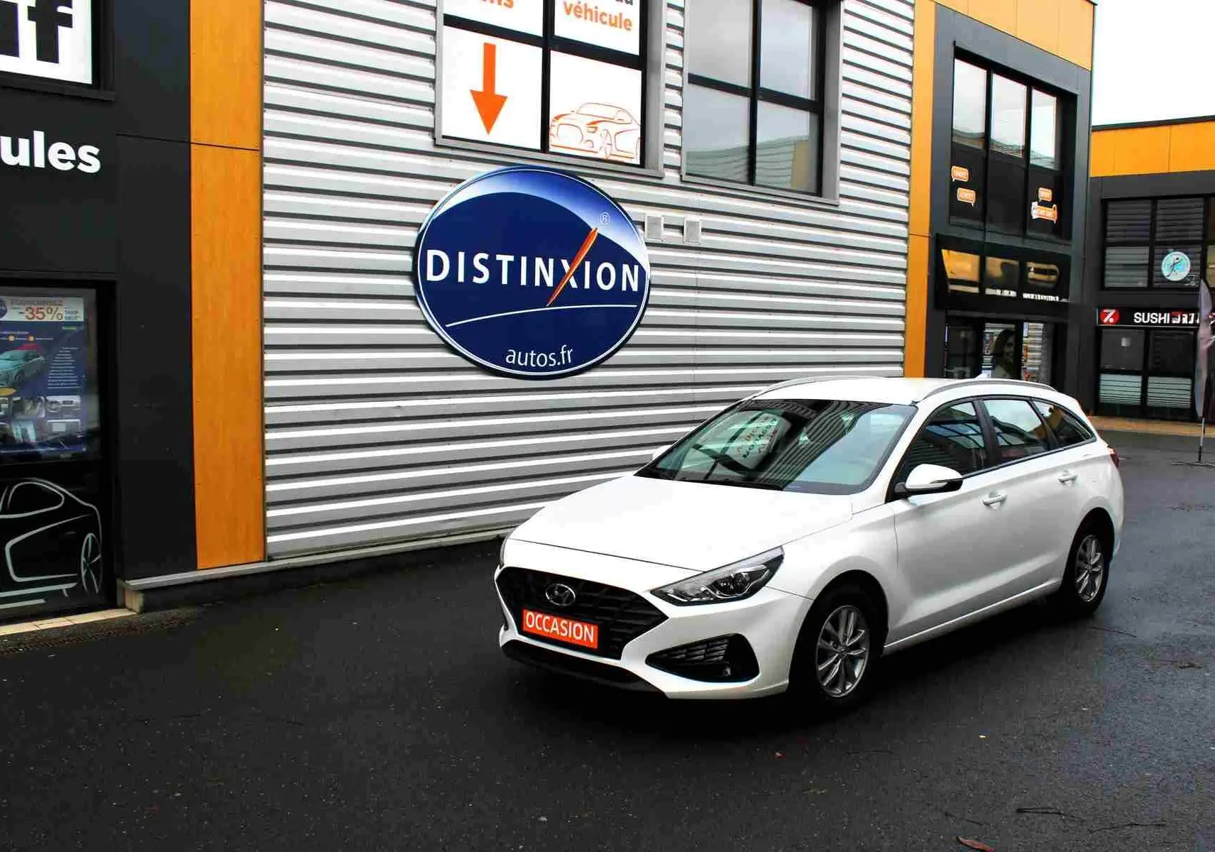 Hyundai i30 SW blanc polaire vue 3/4 avant droit devant un bâtiment avec panneau Distinxion.