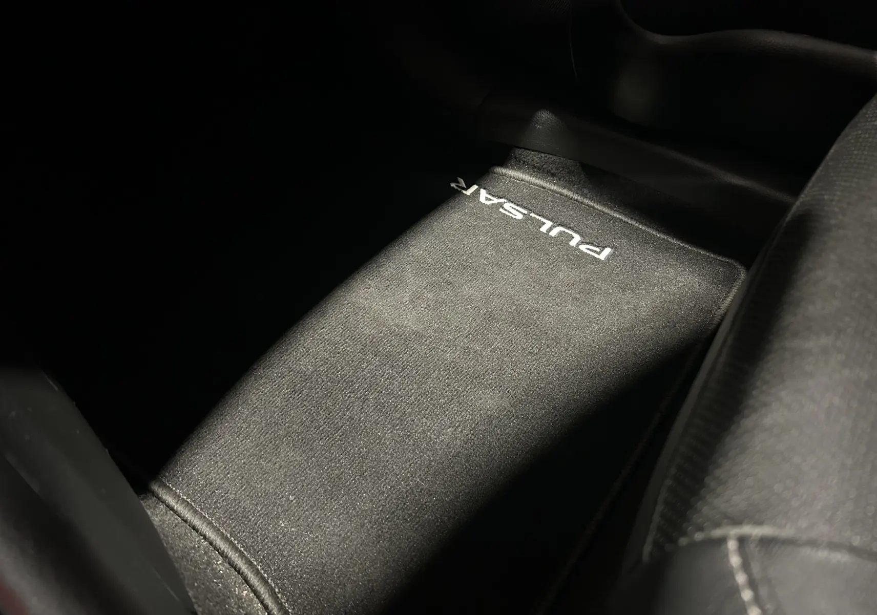 Tapis de sol noir avec inscription "PULSAR" en blanc, vue en plongée sur l'intérieur côté passager.