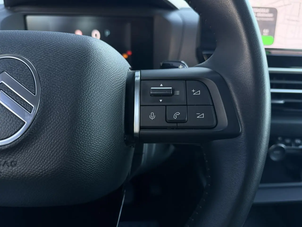 Gros plan sur les commandes du volant en cuir noir d'une Citroën C4 Hybrid 2025 avec écran tactile et navigation visible en arrière-plan.