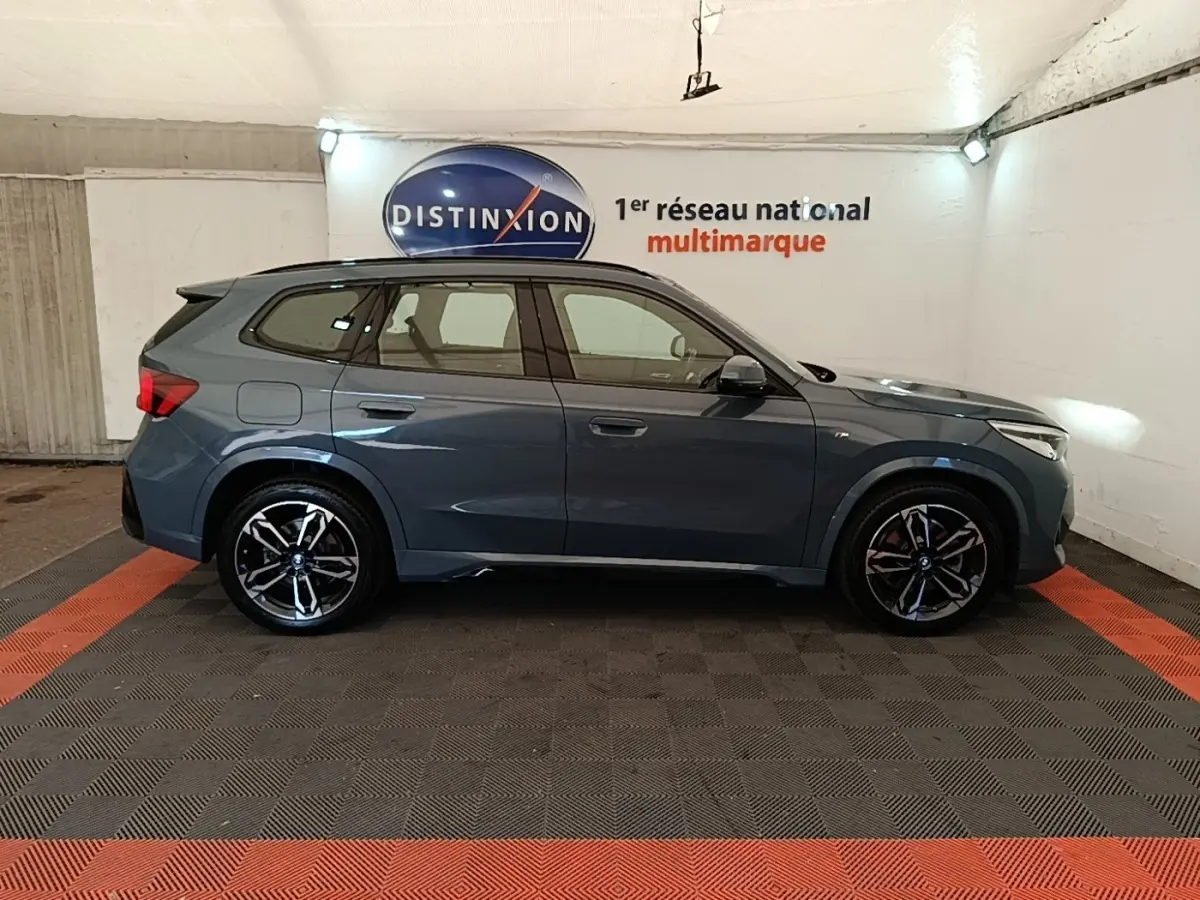 Profil droit d'un BMW X1 xDrive25e M Sport gris foncé, avec jantes alliage et toit spoiler visible en intérieur showroom.
