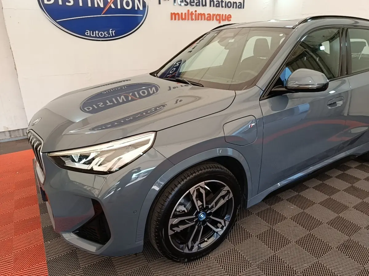 Vue 3/4 avant droit d’un BMW X1 xDrive25e M Sport gris foncé avec jantes noires et phares LED allumés.
