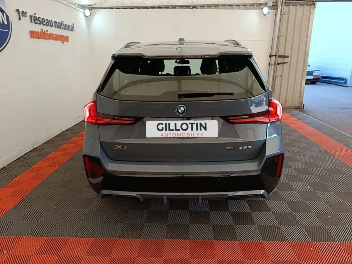 Vue arrière d’un BMW X1 xDrive25e M Sport gris foncé avec feux LED et spoiler de toit visible en intérieur showroom.