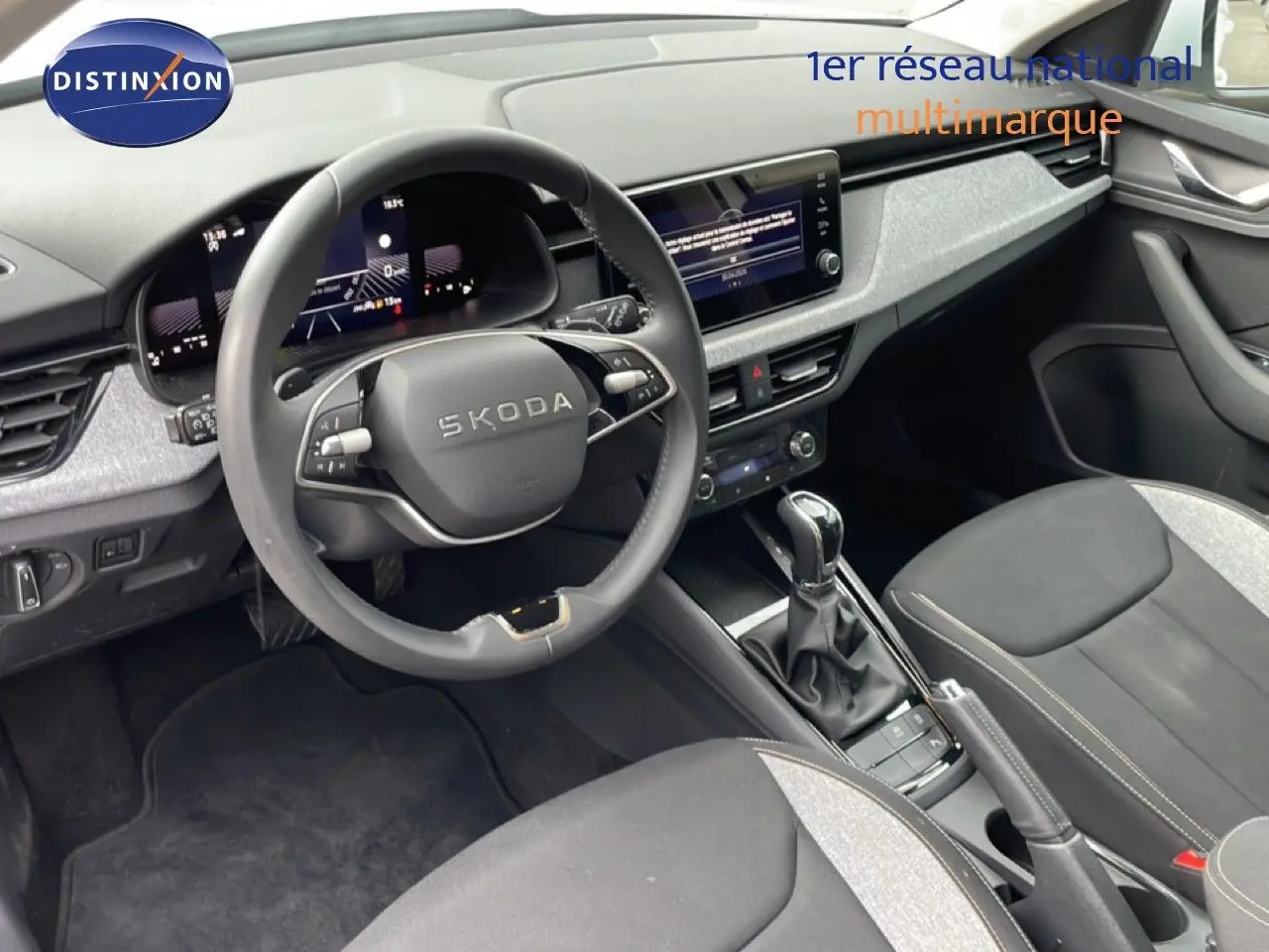Vue intérieure de face du tableau de bord et volant noir du Skoda Kamiq blanc glacier 2025 avec écran tactile et sellerie tissu gris.
